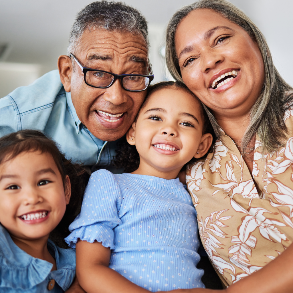 Smiling grandparents hugging two young girls as they pose together in a warm, family setting, conveying joy, closeness, and intergenerational love.